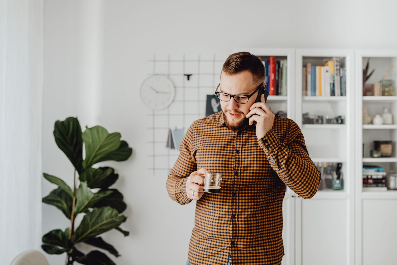 A man in a checkered shirt talks on a phone while holding a glass of coffee