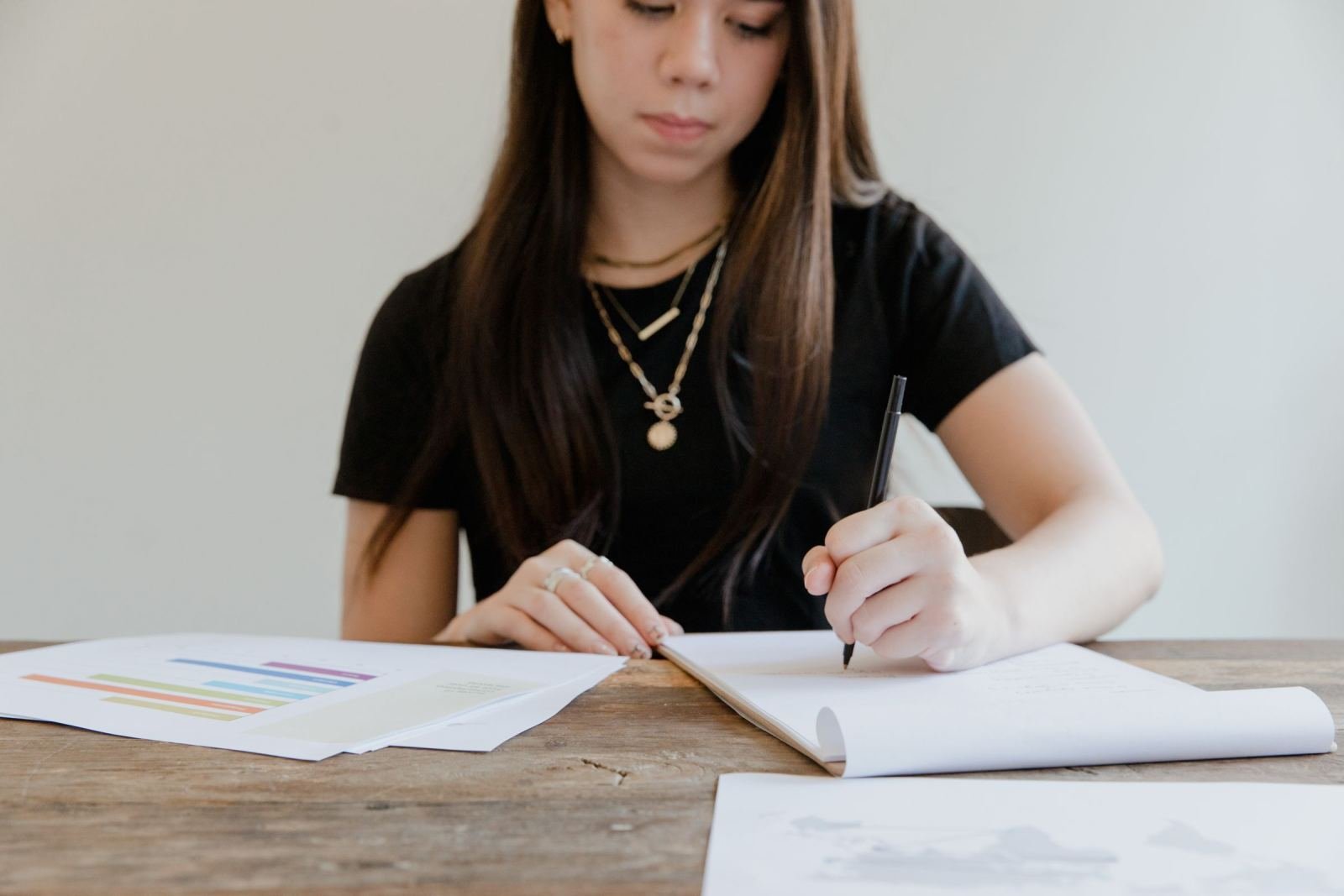 A woman writes in a notebook at a wooden table with papers nearby