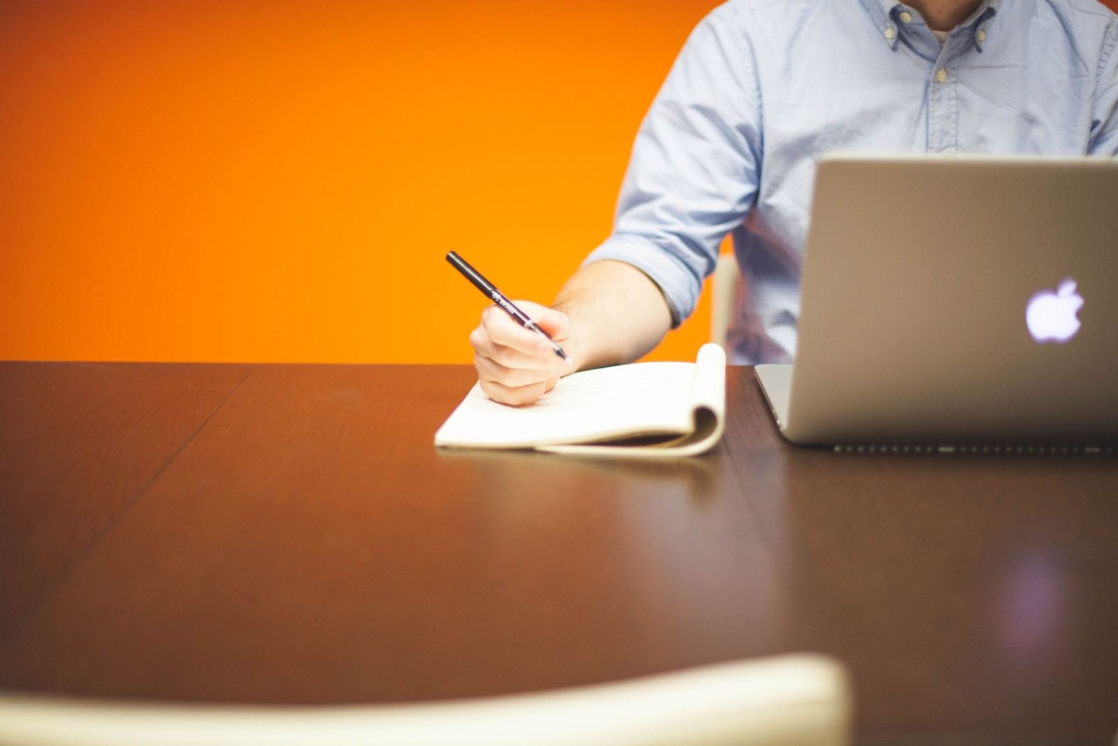 A person writing in a notebook next to an open laptop on a wooden table