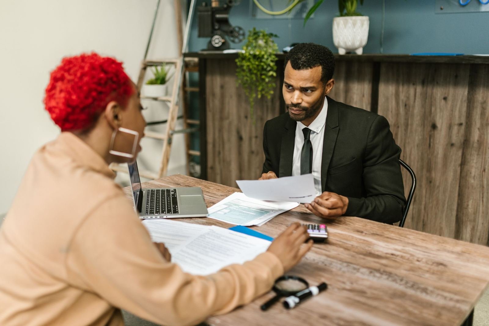 A suited man and a red-haired woman look over documents at a wood table.
