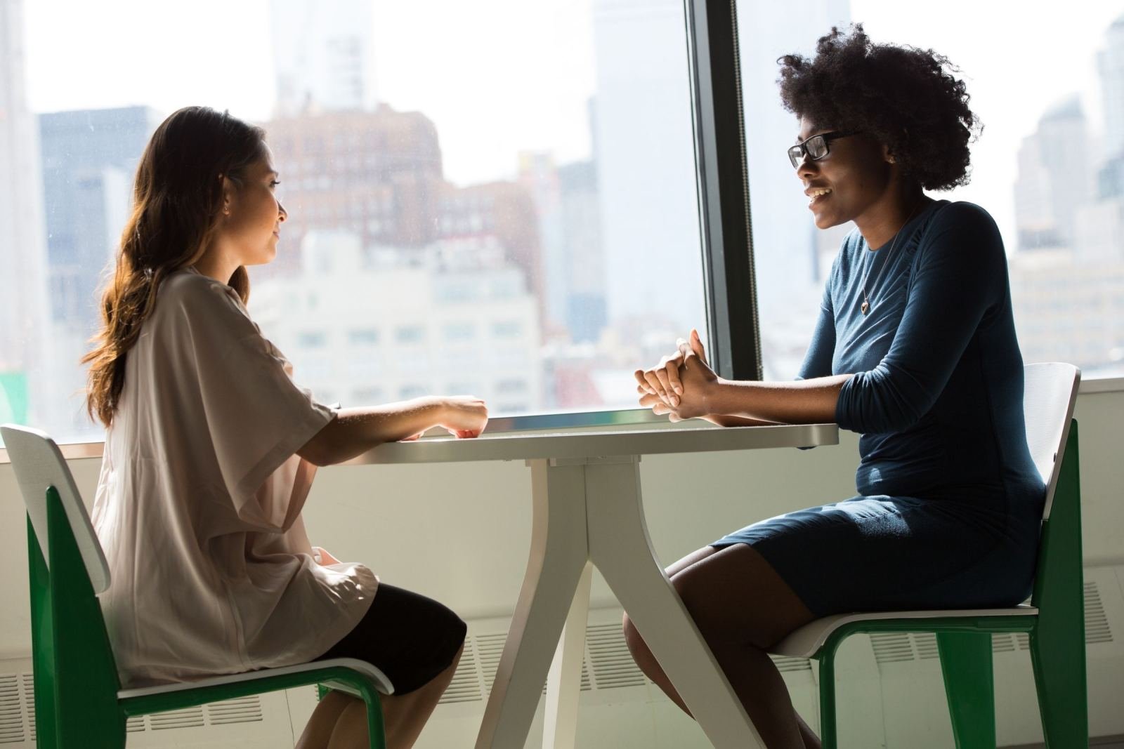 Two women sit at a white table in a bright office talking