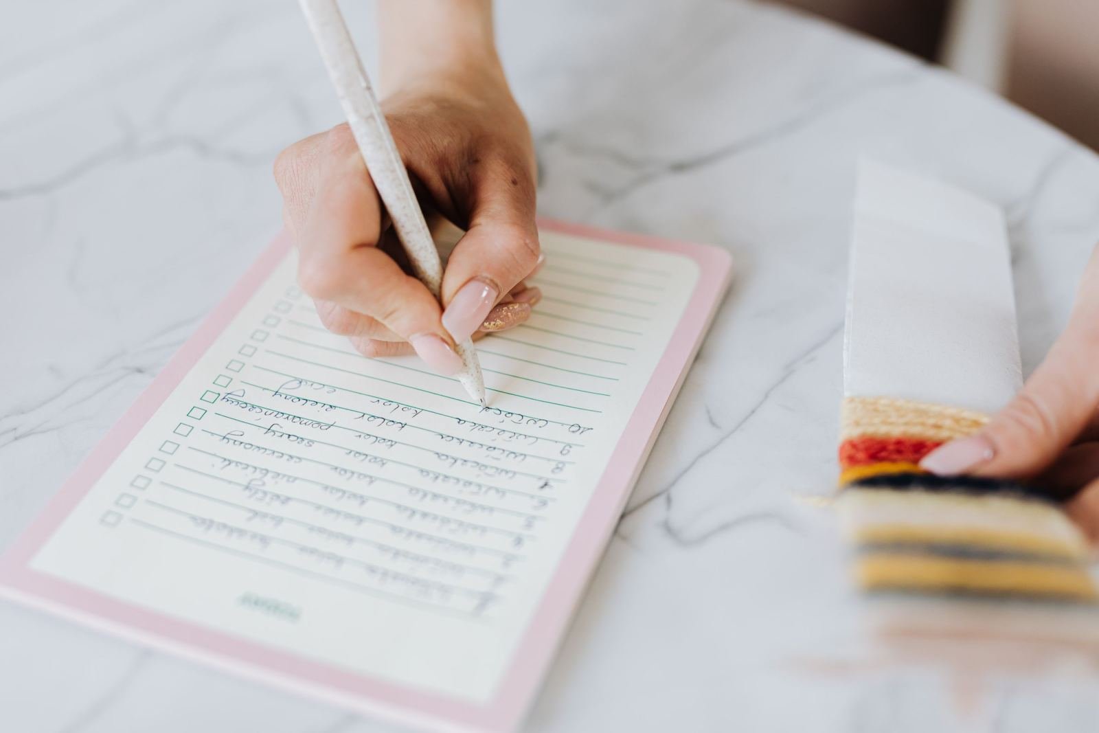 A person writes on a checklist in a pink notebook on a marble table