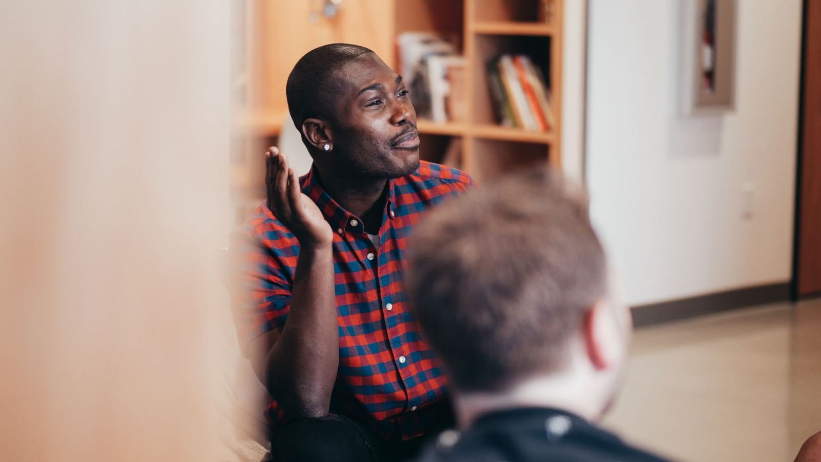 A man wearing a plaid shirt uses his hands while he talks to someone else in an office.