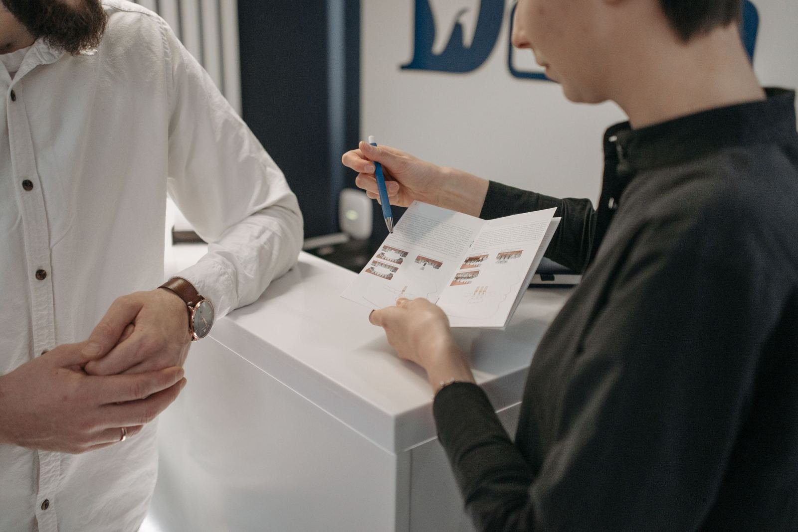 A person points to a brochure while talking to a man at a white counter