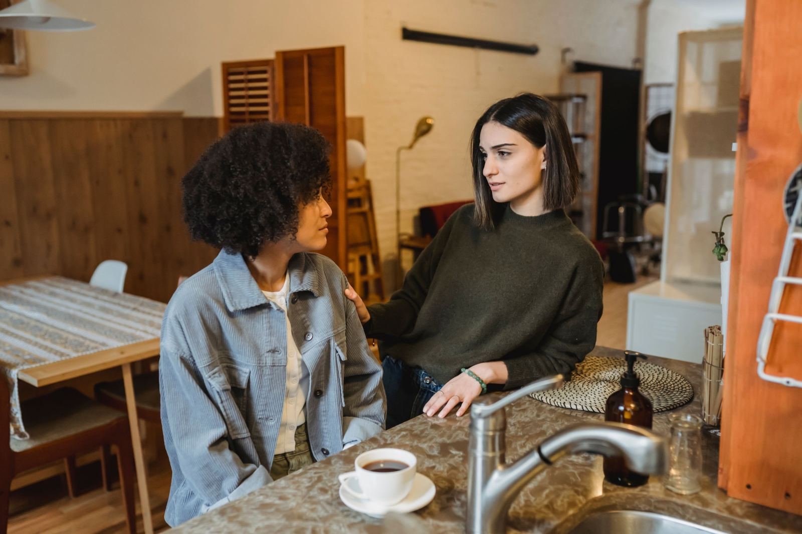 Two women talk in a kitchen, one with a supportive hand on the other's shoulder