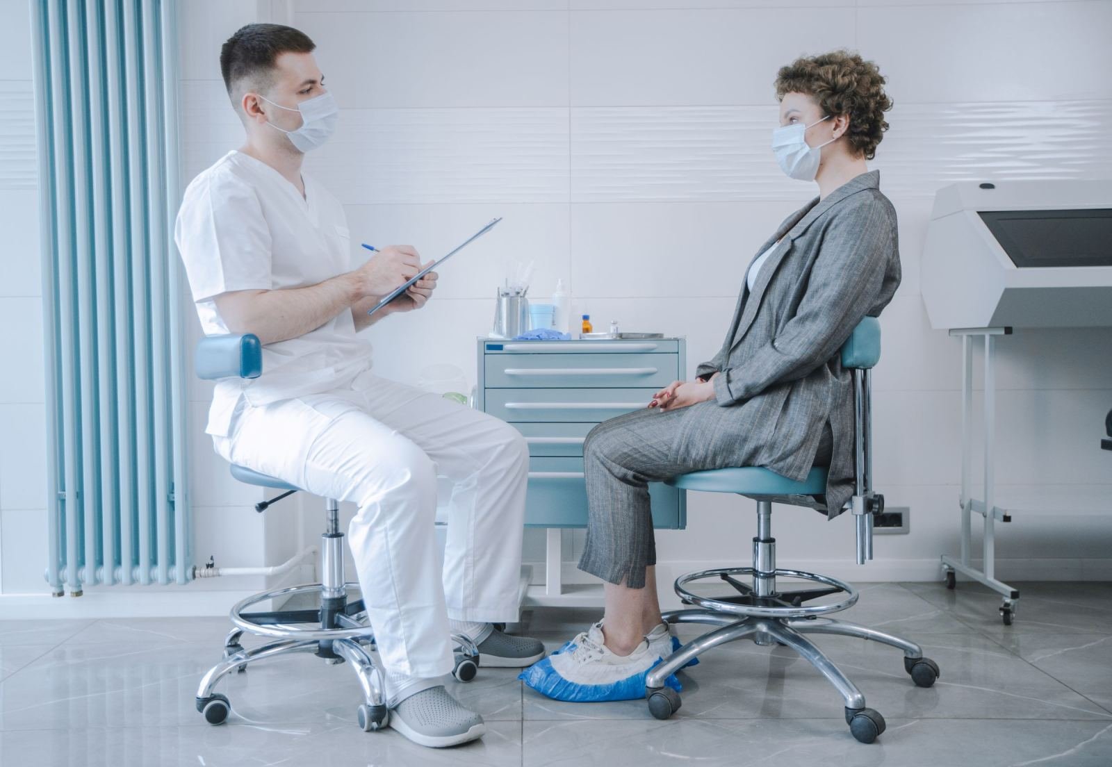 A masked patient in a suit and a masked doctor in scrubs chat in a clinic.