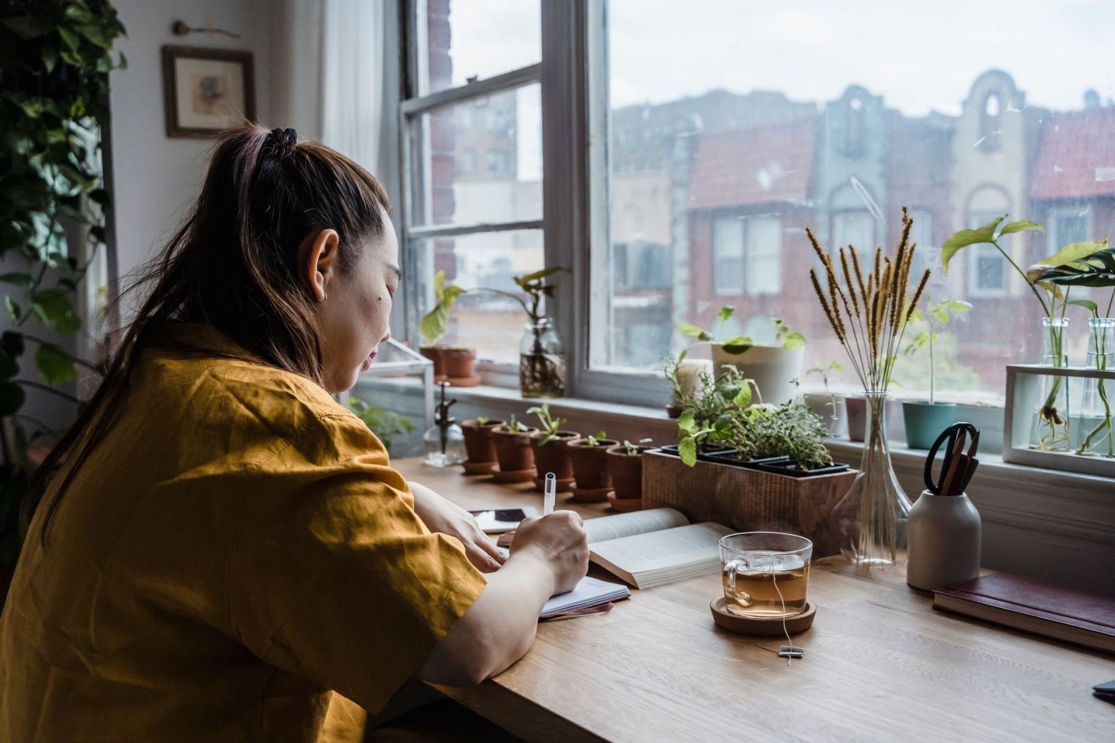 A person in a yellow shirt writes in a journal at a table by a window