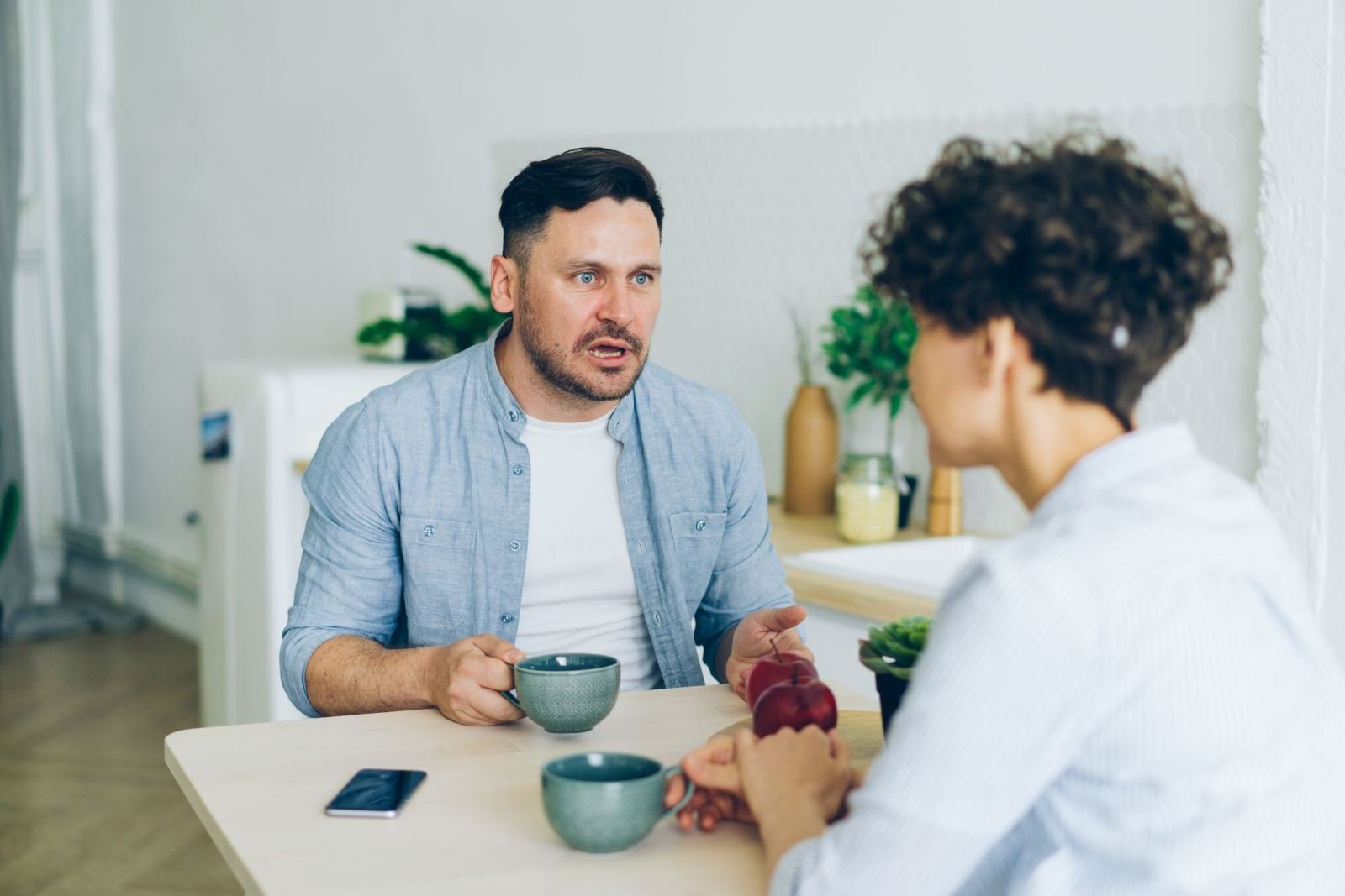 A man and woman sit at a table talking over mugs of coffee