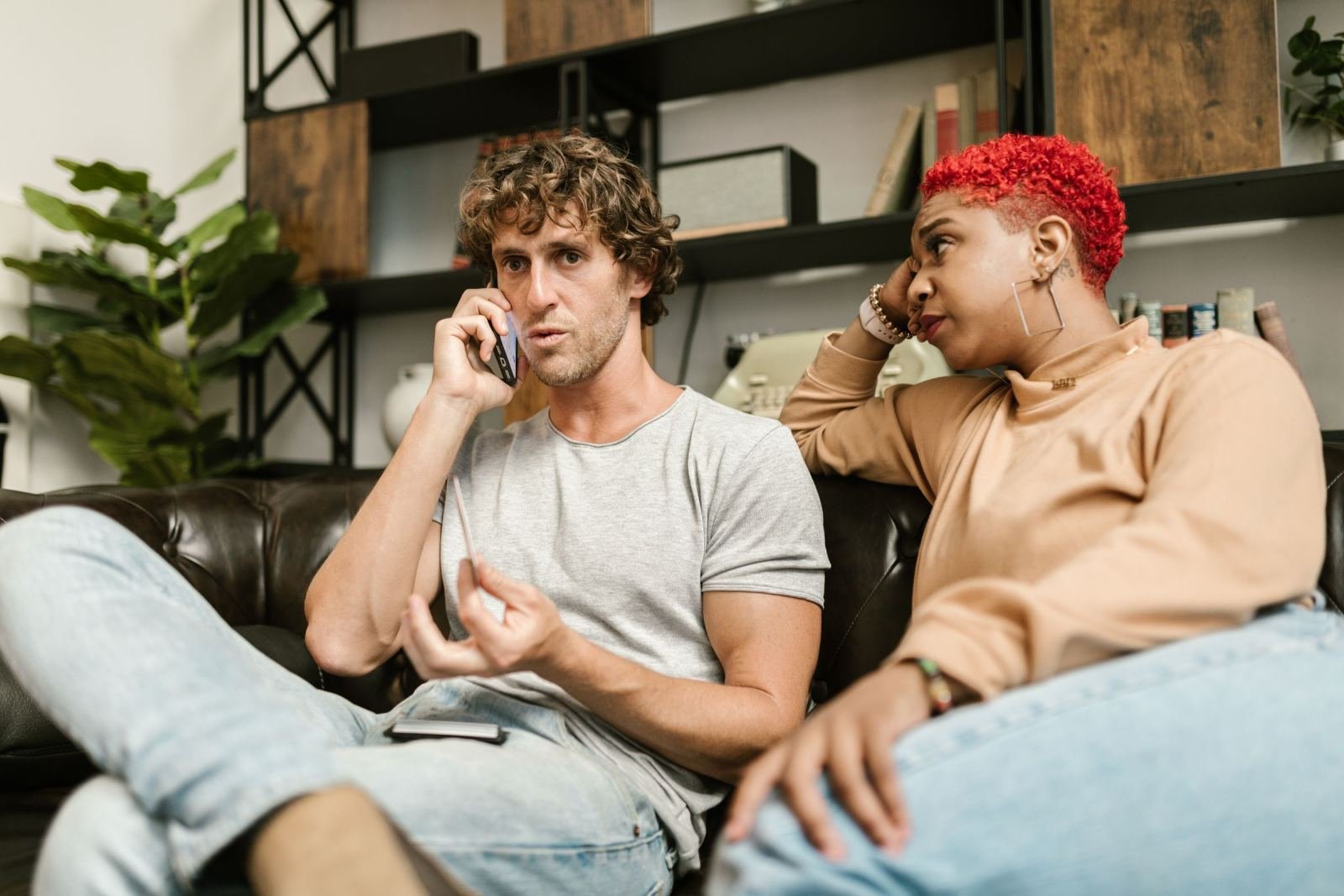 A man makes a phone call while a woman with red hair sits beside him on a leather sofa