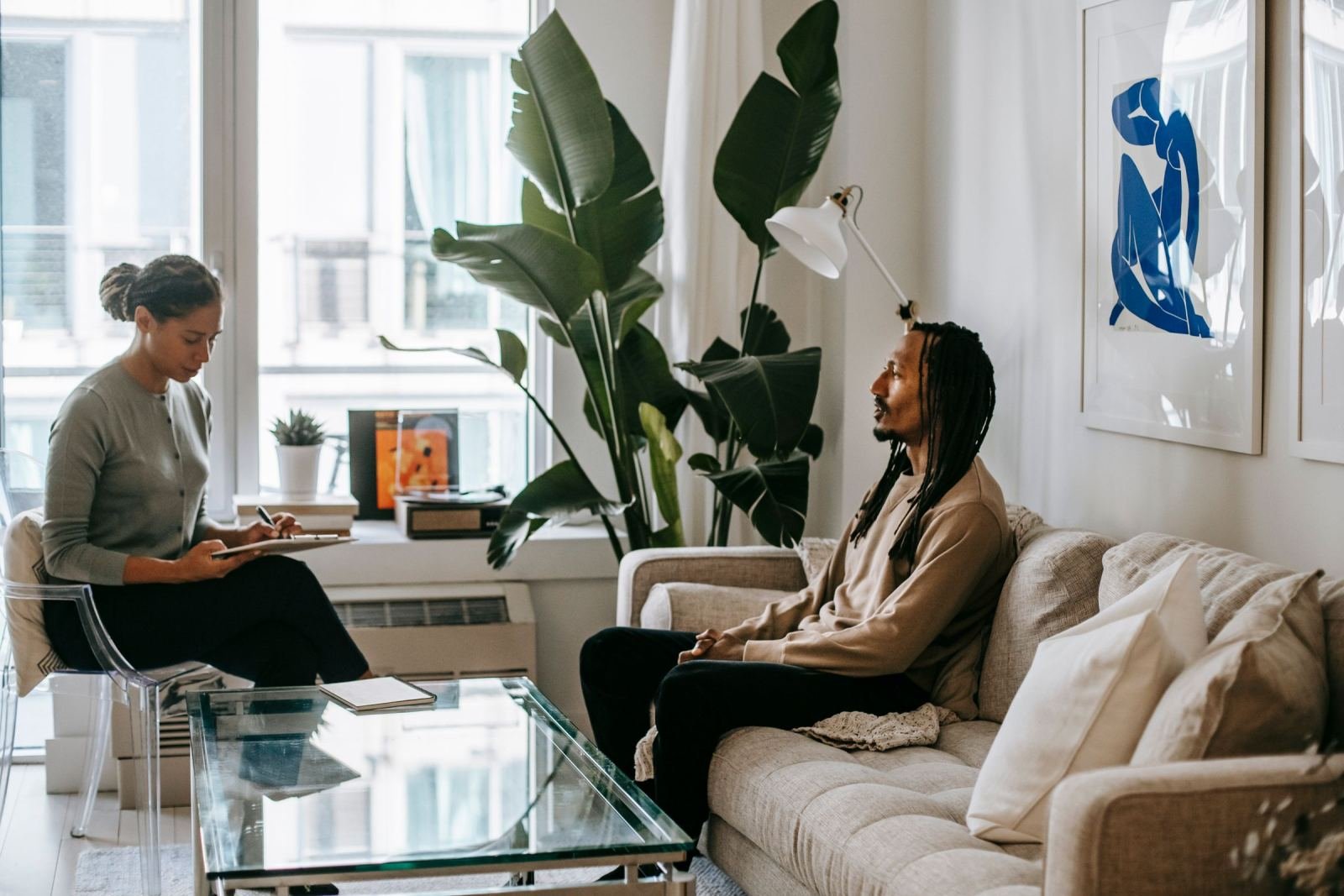 A woman with a notebook talks with a man sitting on a sofa in a bright room