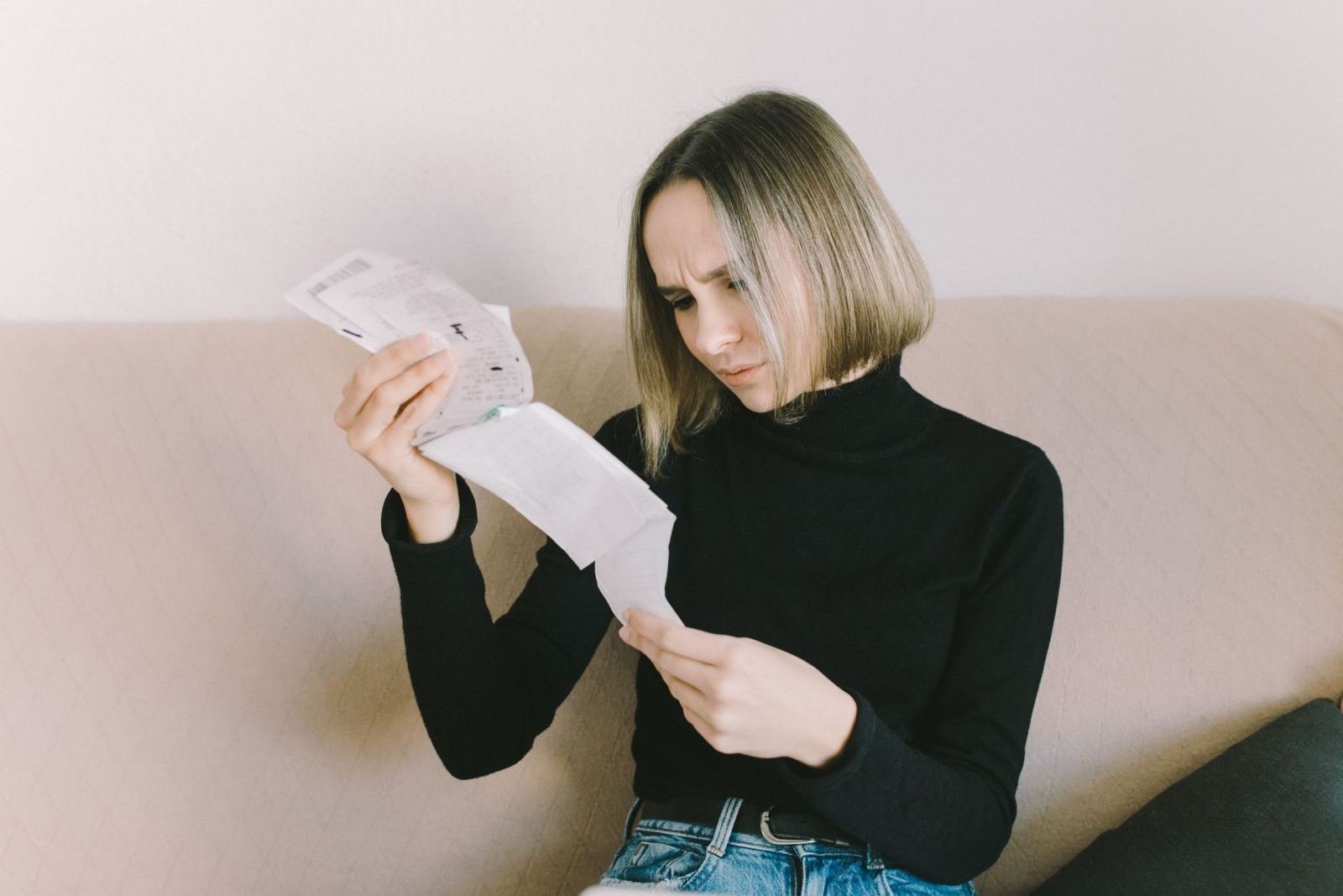A woman sits on a couch looking at a piece of paper with a concerned expression