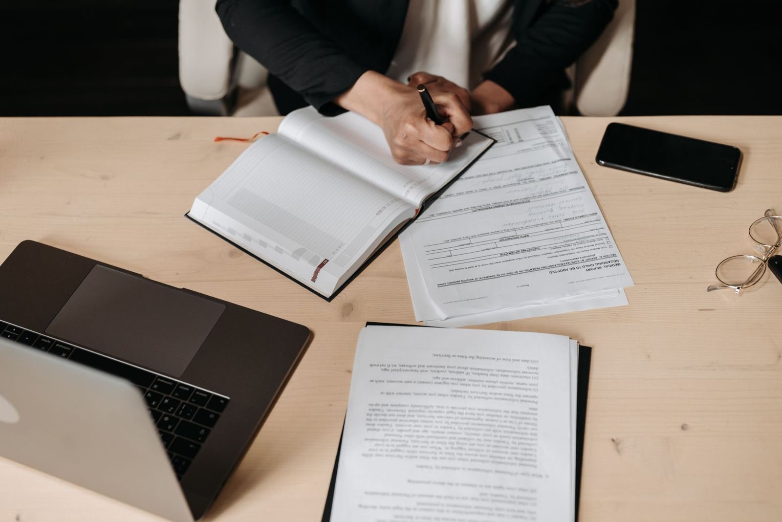A person writes in a notebook next to insurance papers, a laptop, and a phone