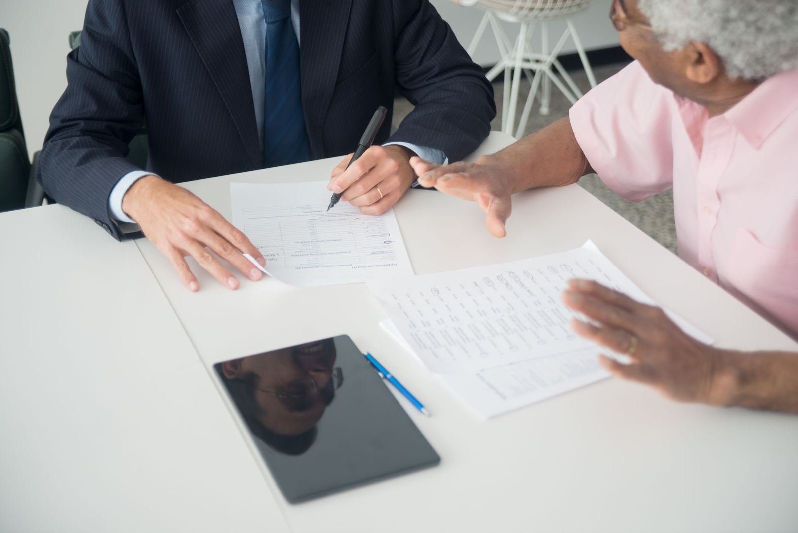 An older man and a professional in a suit review documents at a white desk