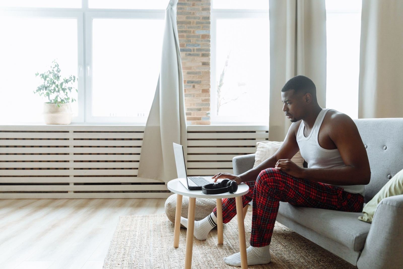 A man in pajama pants sits on a sofa using a laptop on a small table
