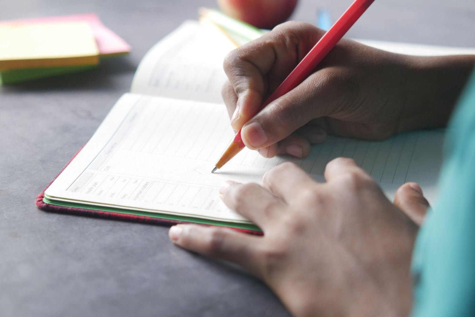 A person's hands writing in a planner with a red pencil
