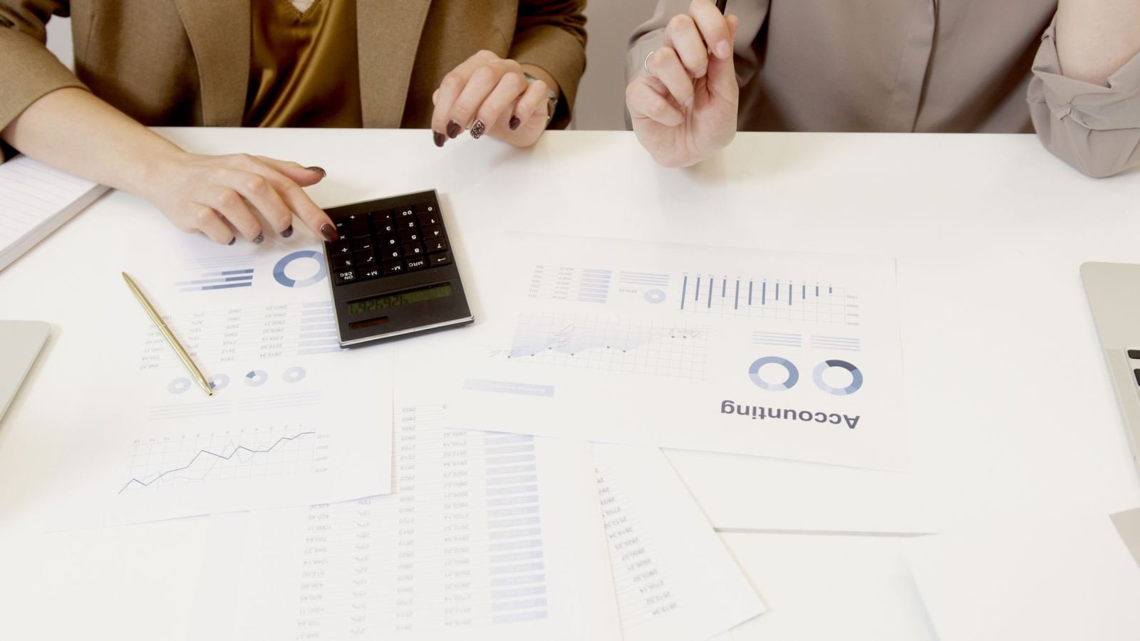 Two people review accounting documents and use a calculator at a white table