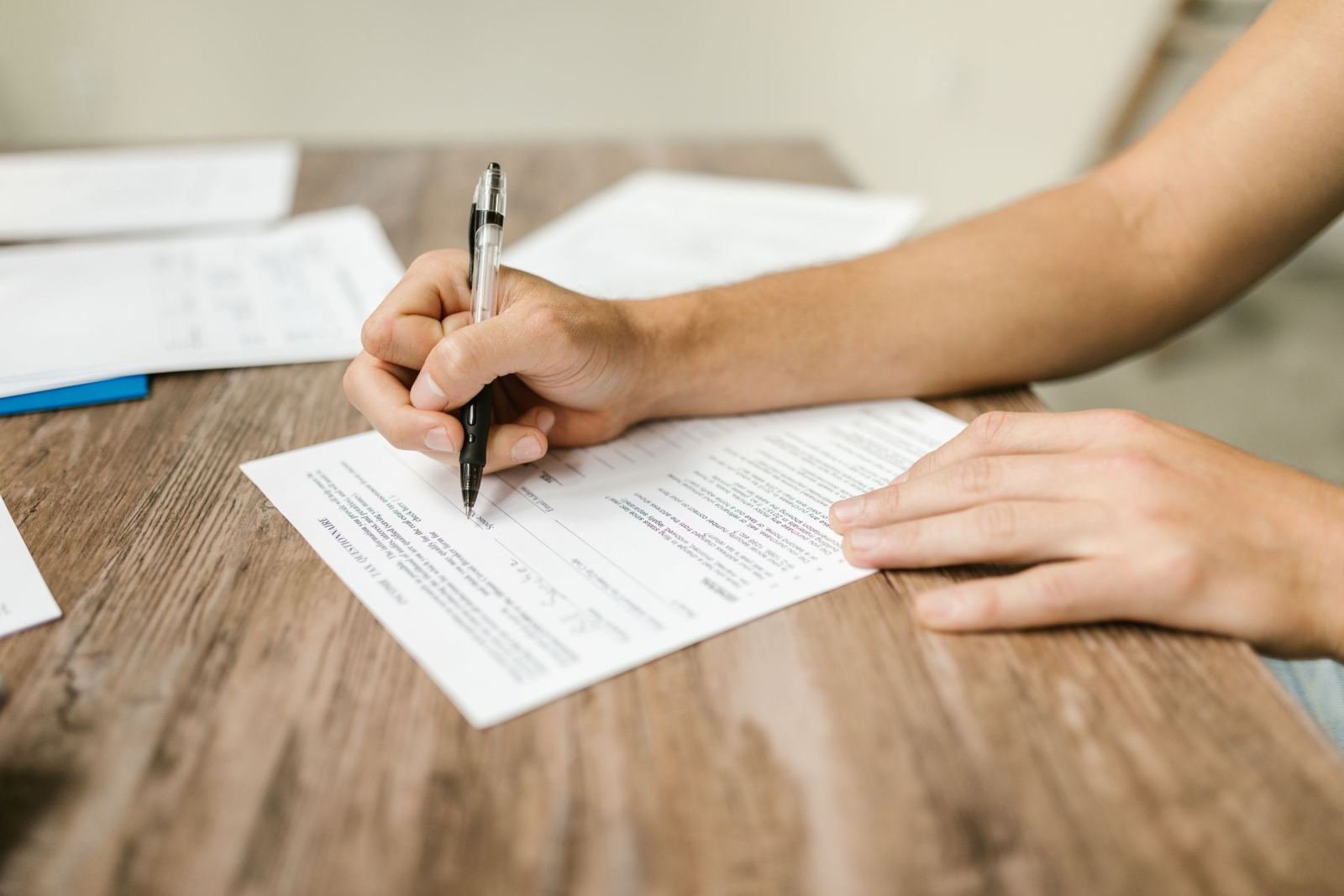 A person filling out a white document with a black pen on a wooden table