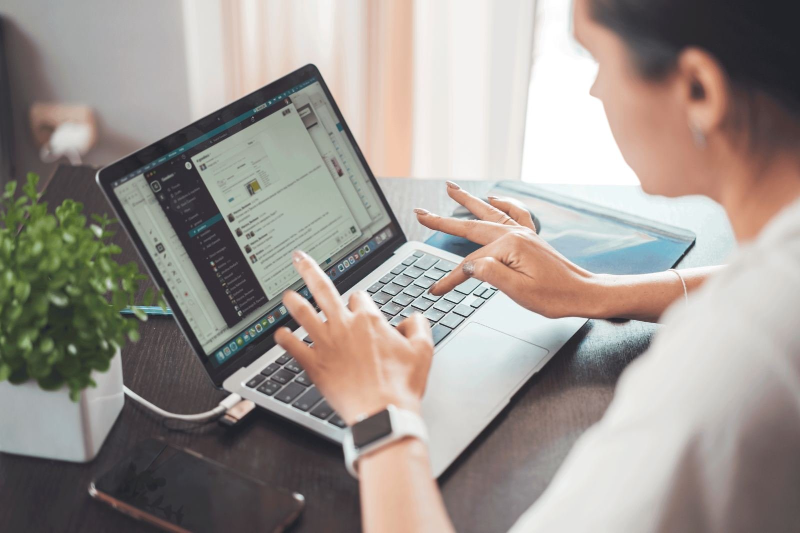 A person uses a laptop and smartphone at a desk with a small plant
