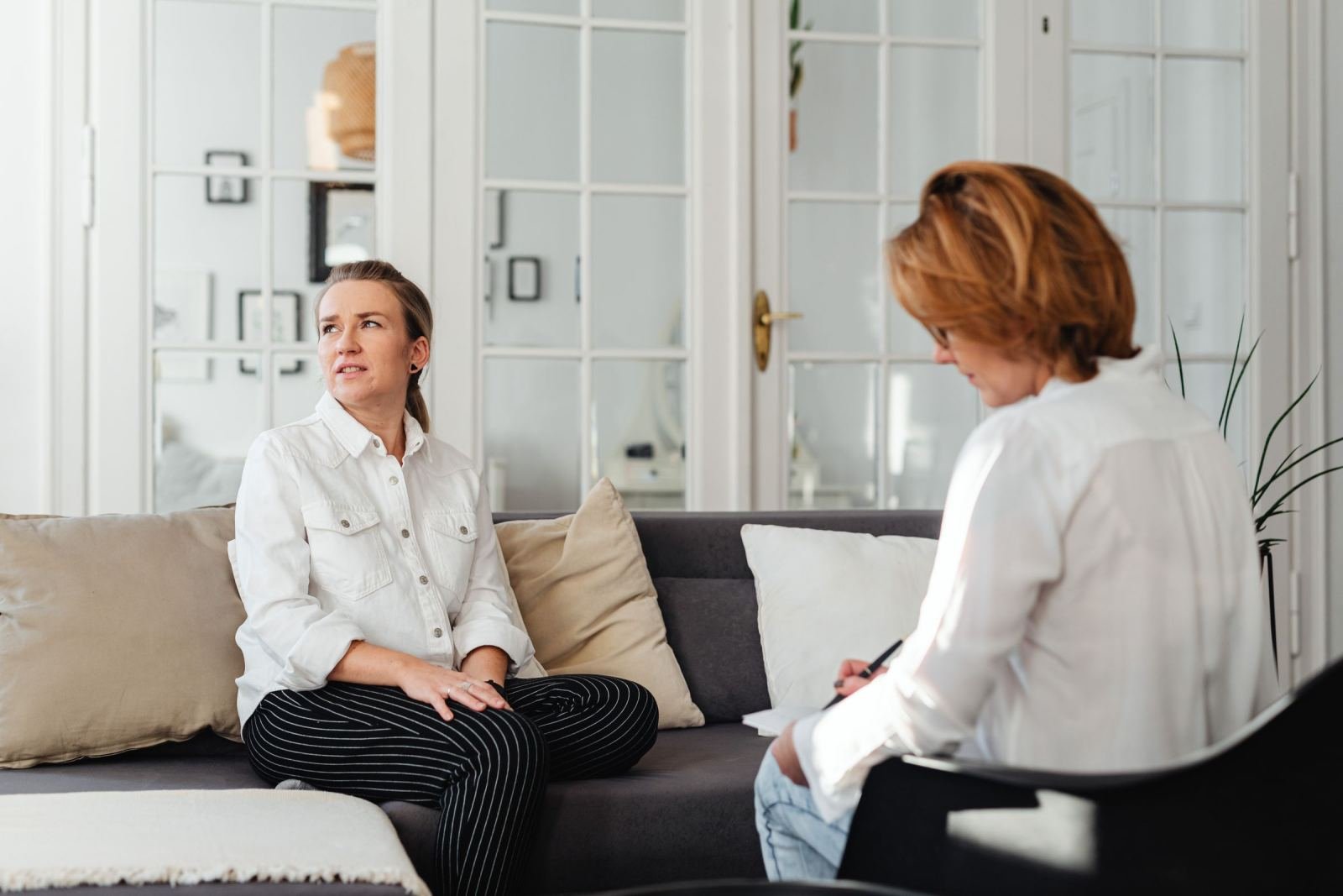 Two women sit and talk in a bright office with a gray sofa