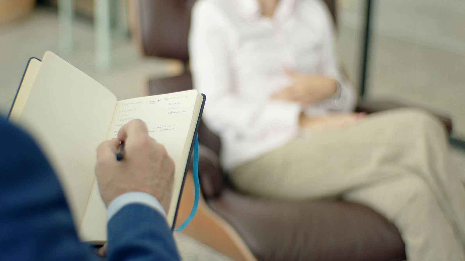 A person in a suit takes notes in a notebook while a client sits in a chair