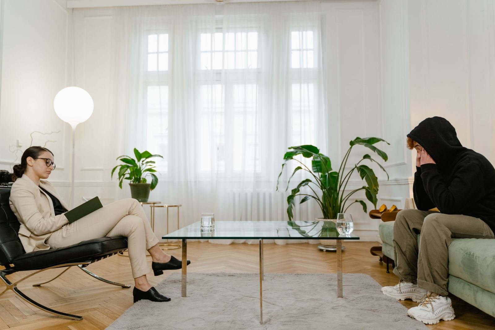 A counselor in a beige suit talks with a person in a black hoodie in a bright office