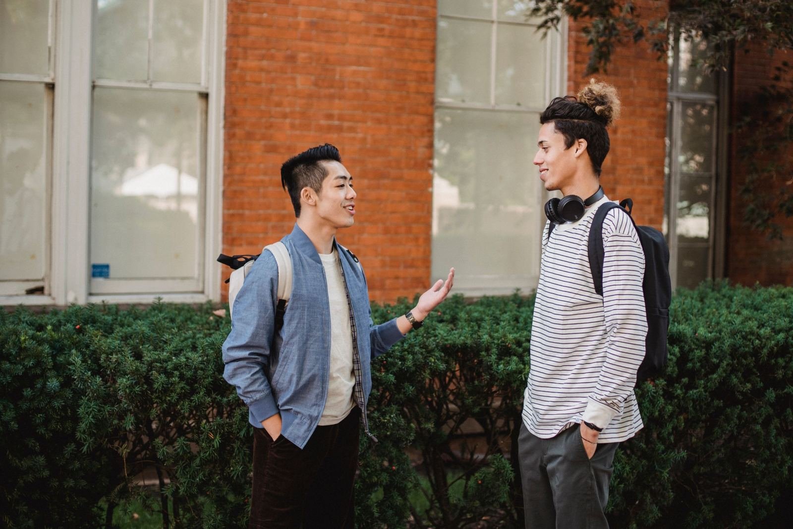 Two young men with backpacks talk in front of a brick building and green hedges