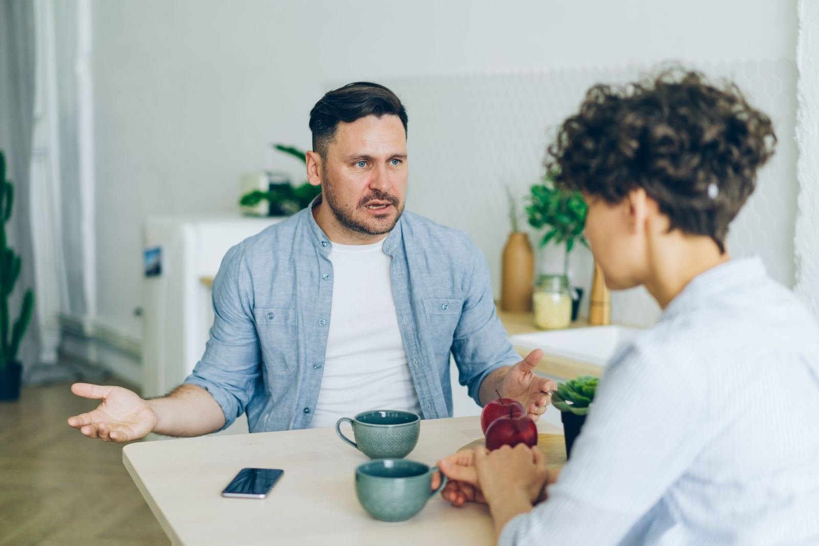 A man gestures while talking to a woman at a kitchen table with mugs and apples