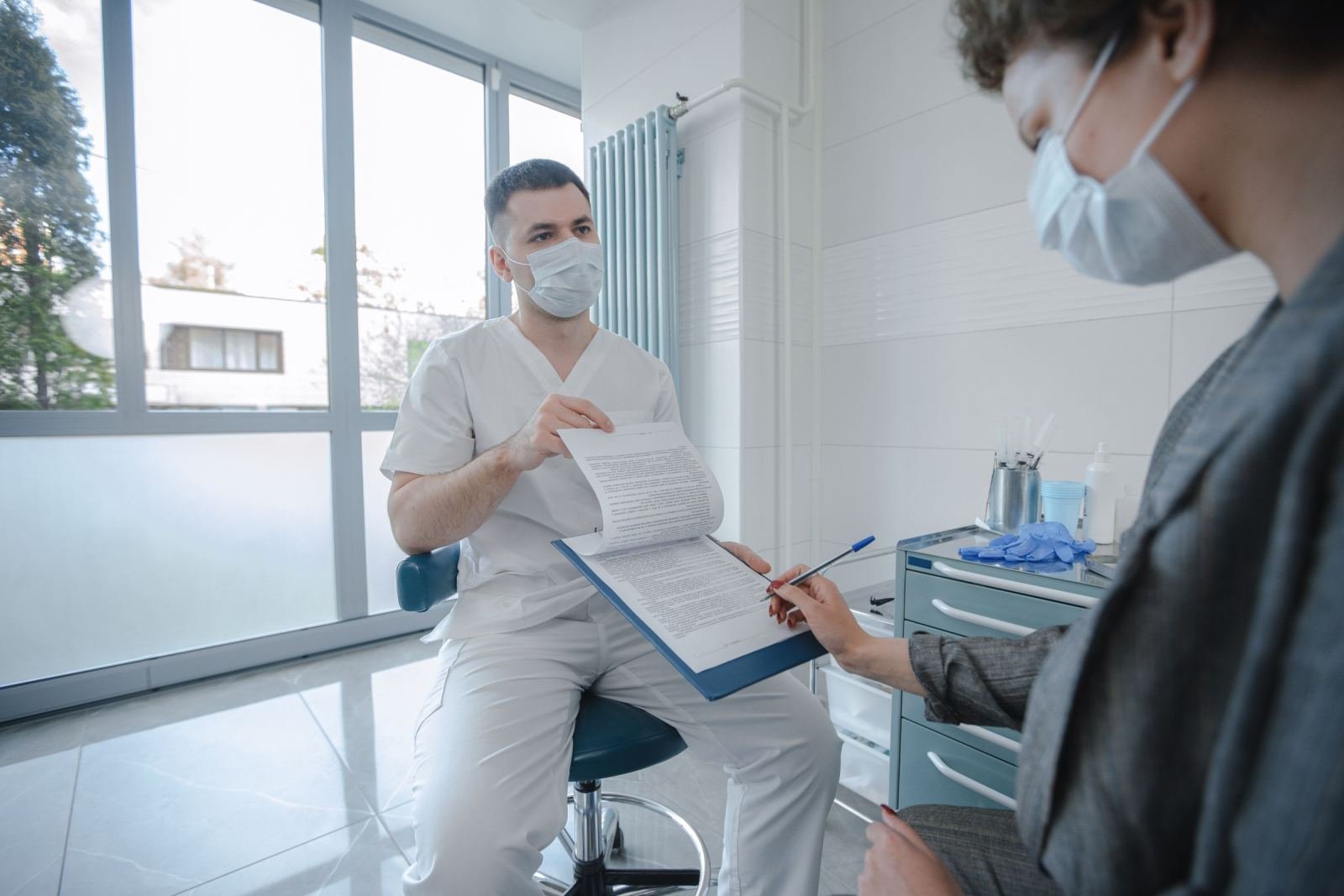 A medical professional in scrubs and a mask reviews a document with a patient