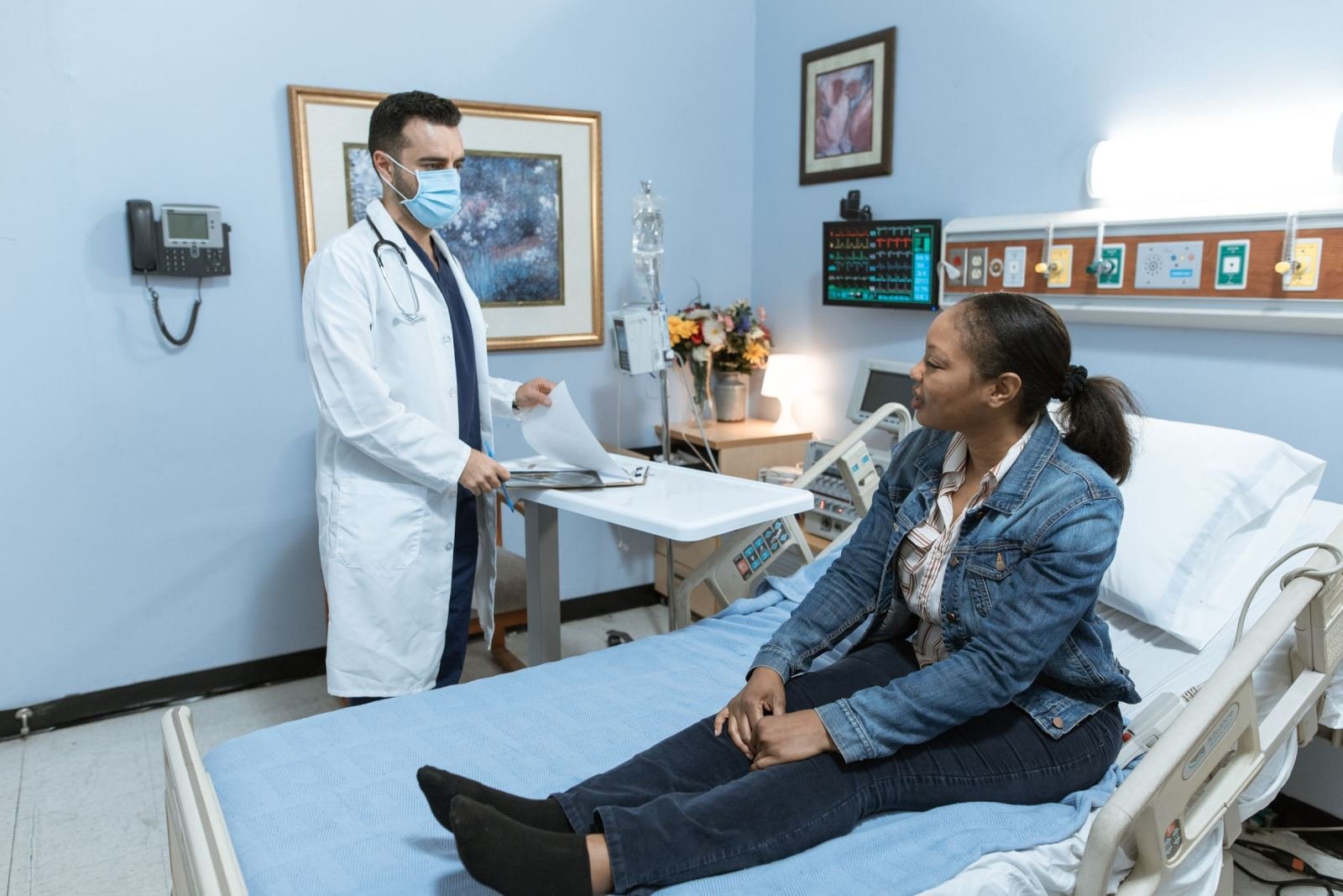 A doctor in a face mask talks to a woman sitting in a hospital bed