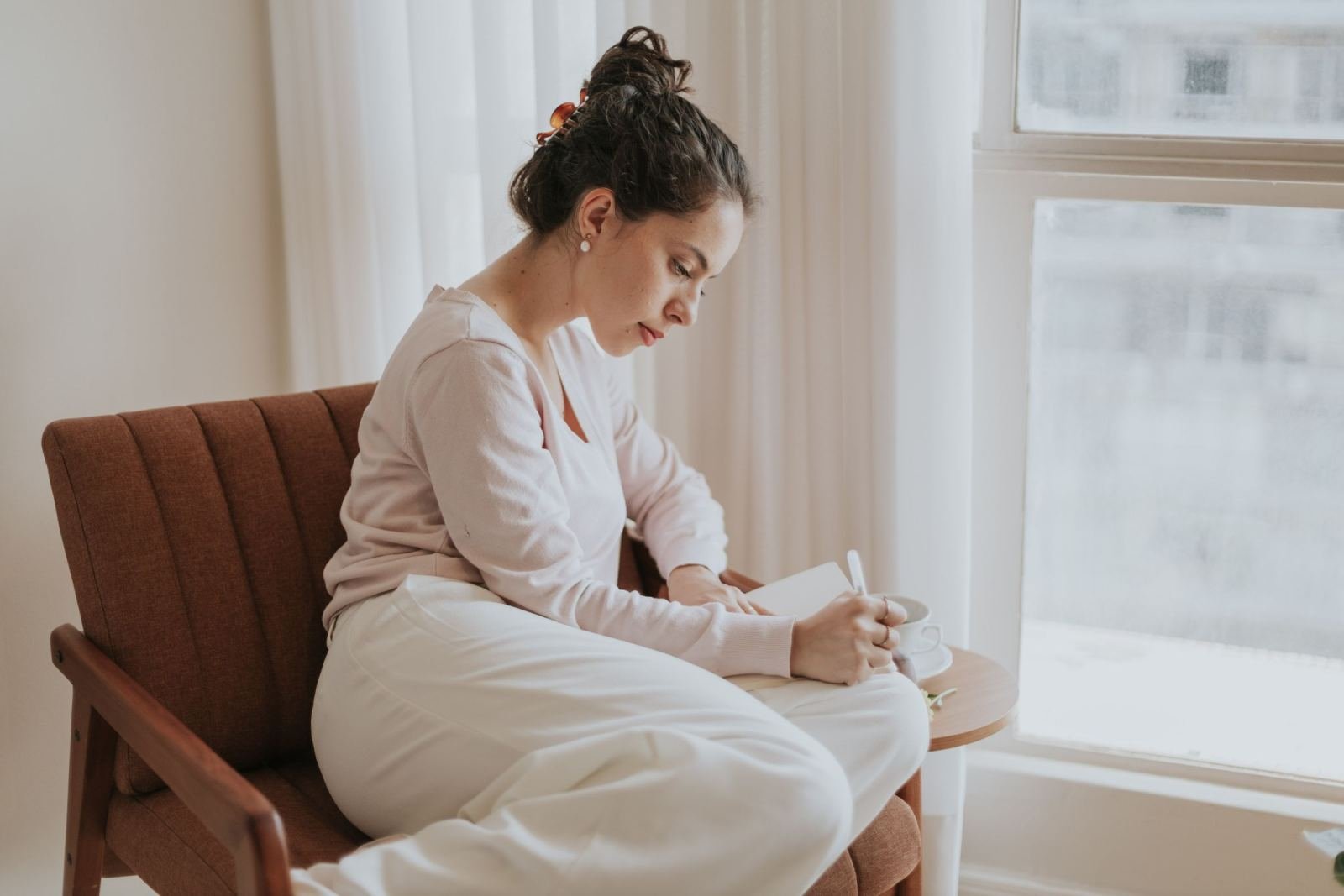 A woman sits near a window in an armchair. She writes in a journal while a cup of tea rests beside her.