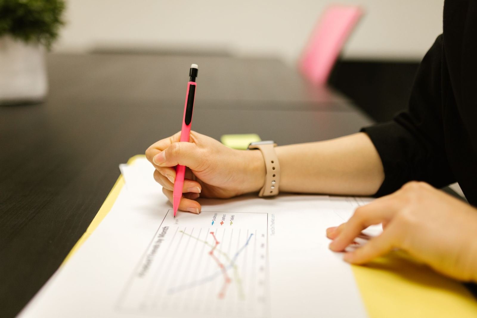 A person uses a pink pen to review a document with line graphs on a dark desk