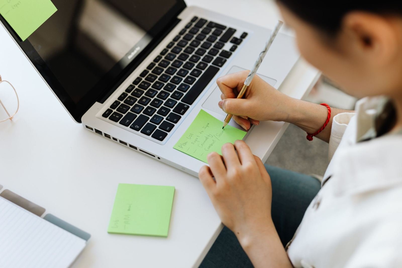A person writing on a green sticky note next to a laptop on a white desk