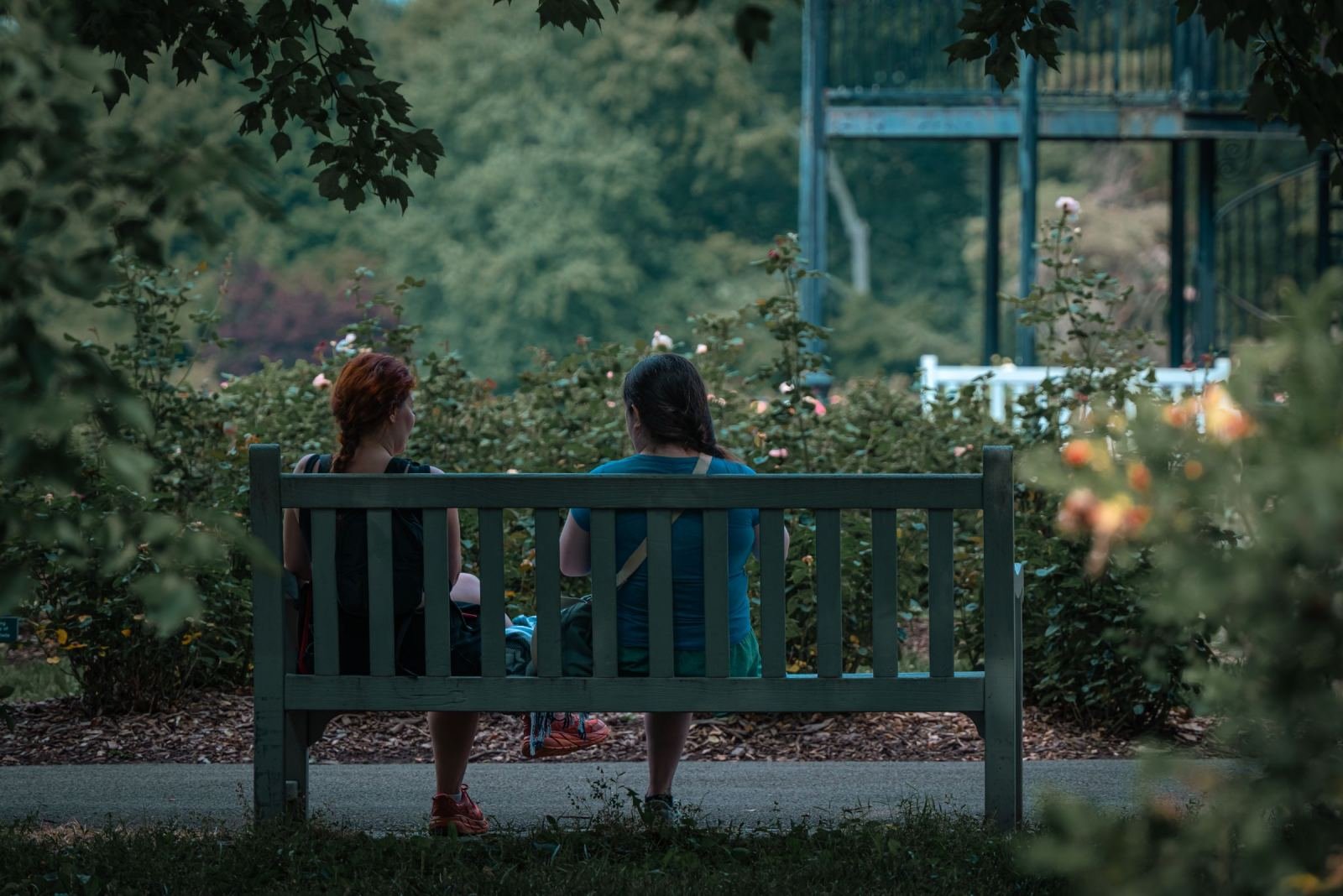 Two women sit side by side on a wooden bench in a garden