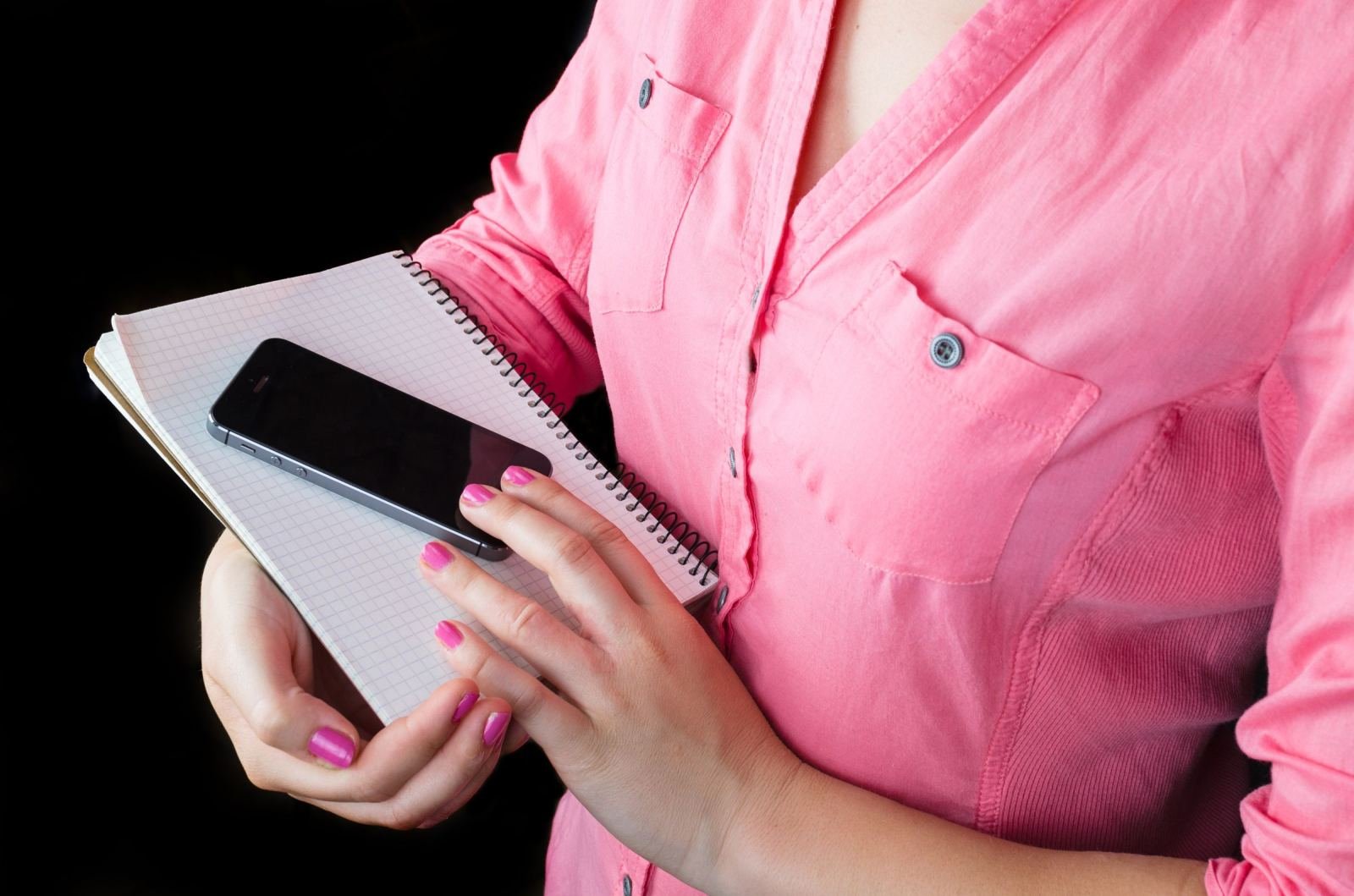 A person in a pink shirt holding a smartphone and a spiral notebook