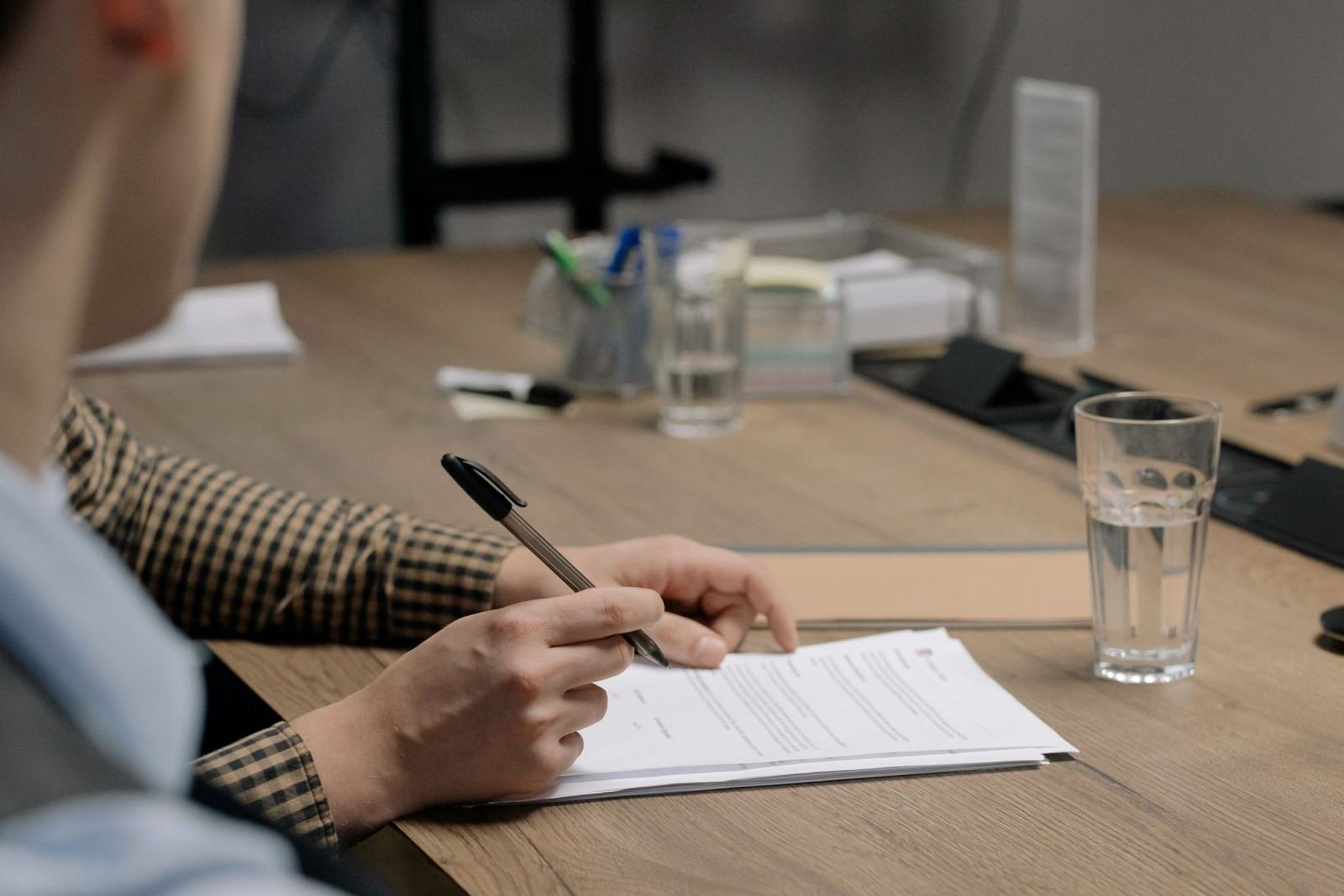 A person in a checkered shirt holds a pen over a document on a wooden desk