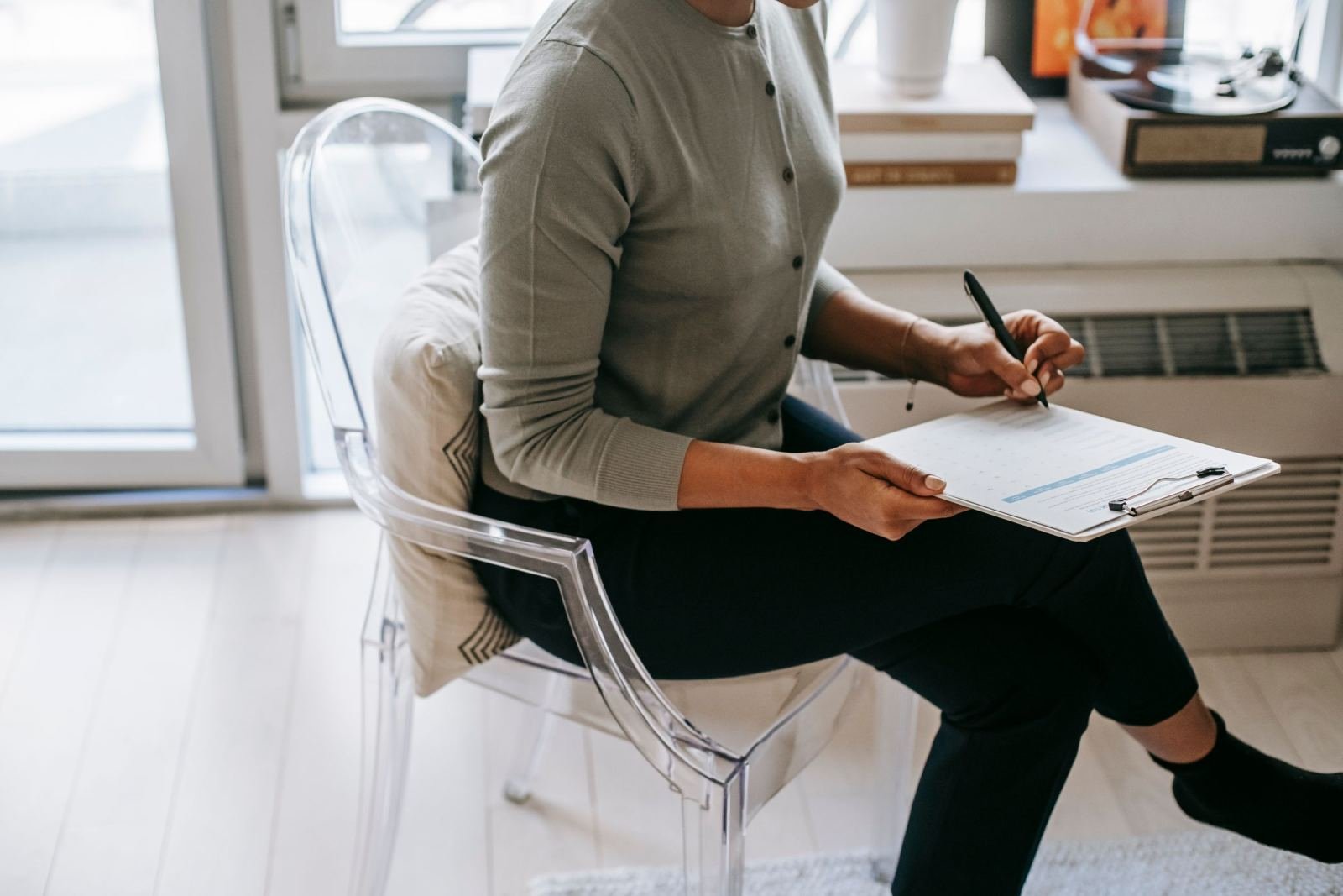 A person in a grey cardigan writes on a clipboard while sitting in a clear chair