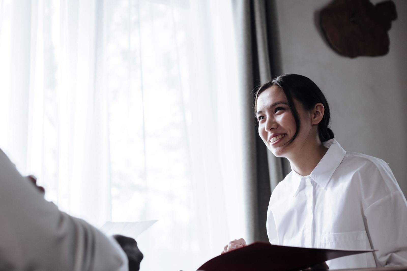 A smiling woman in a white shirt sits at a table talking with another person
