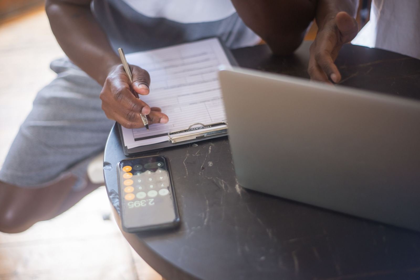 A person writing on a clipboard next to a laptop and a smartphone