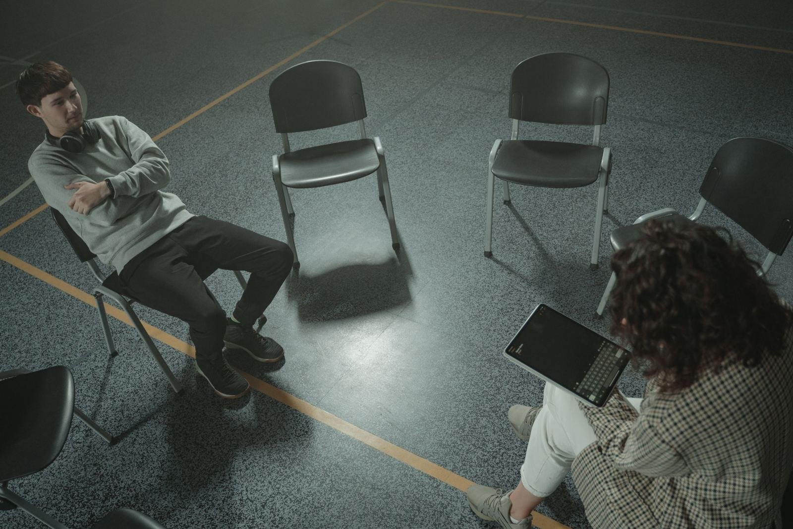 A man sits in a circle of chairs facing a woman holding a tablet