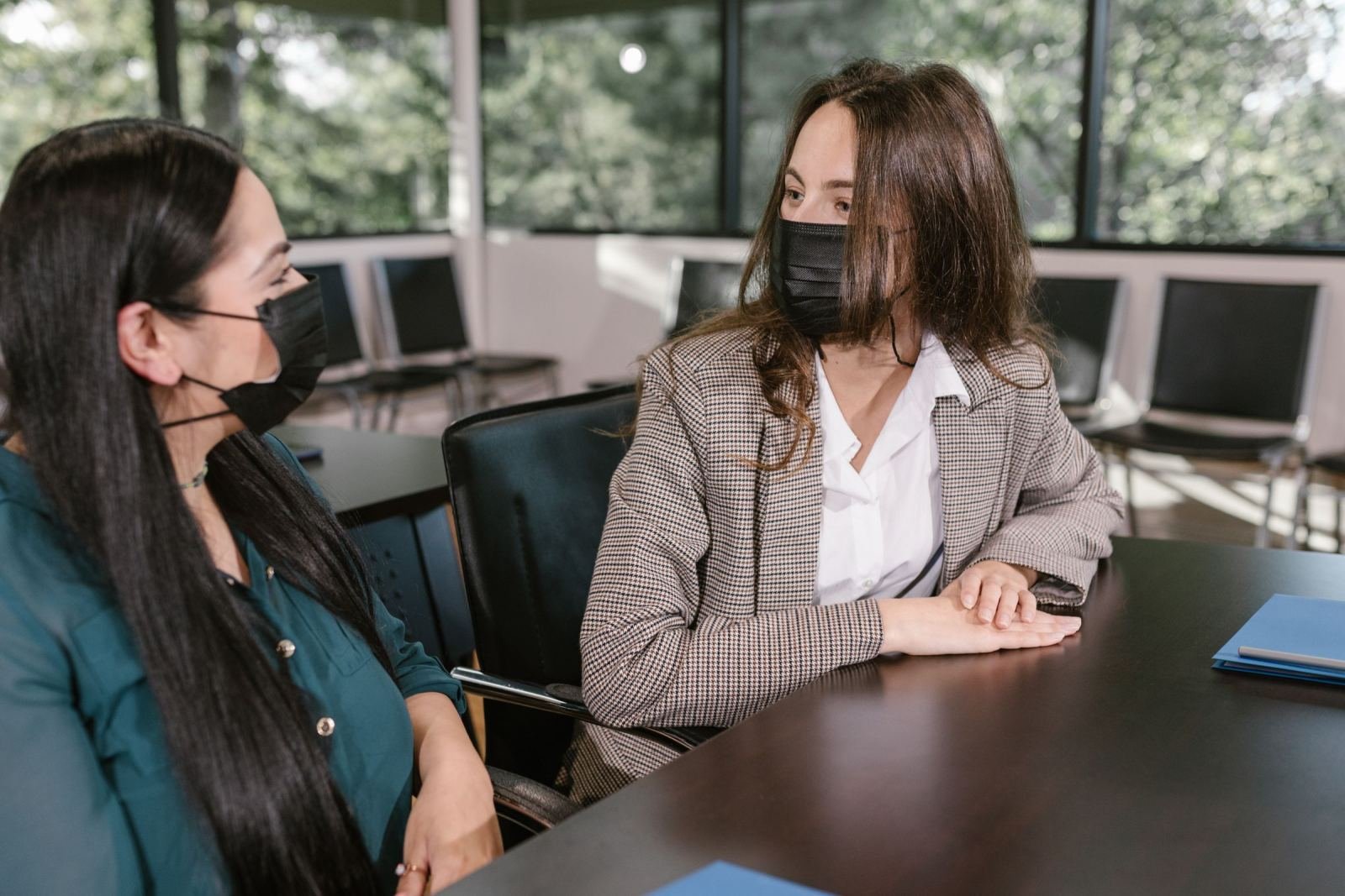 Two women wearing face masks talk across a table in an office