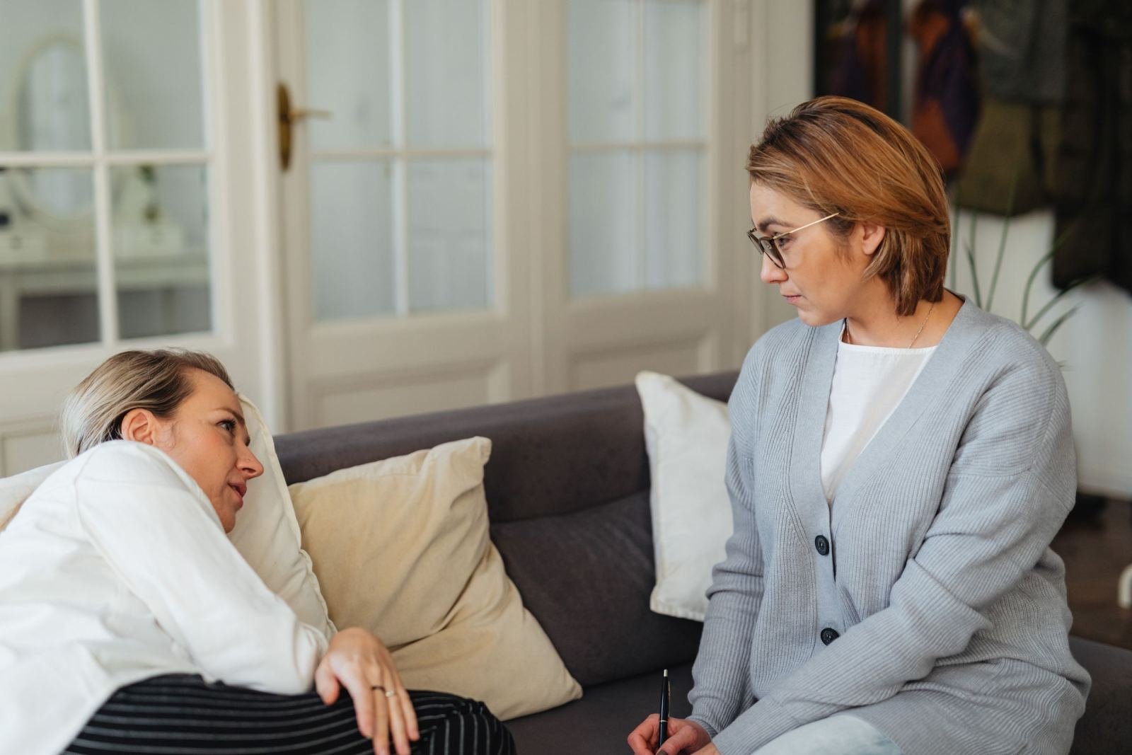 In a sunlit room, one woman sits on a couch while the other holds a pen.