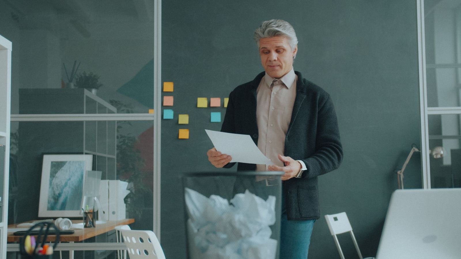 A man in a blazer looks at a piece of paper next to a wastebasket of crumpled paper
