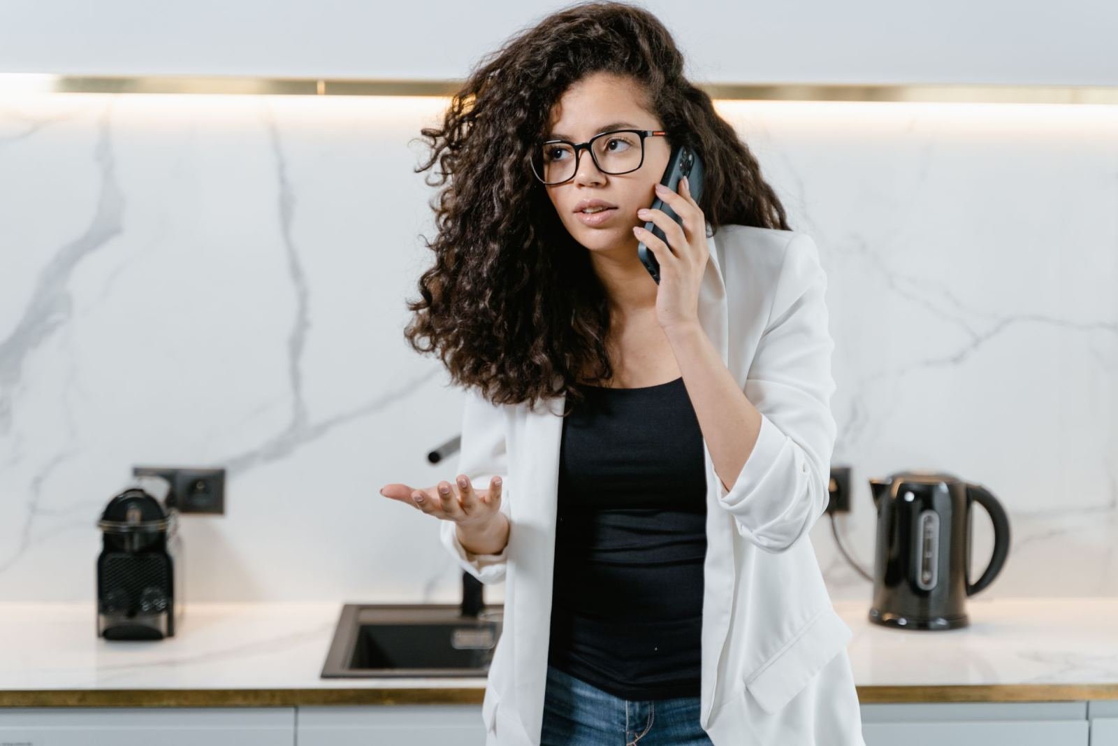 A woman with curly hair and glasses talks on a phone in a kitchen