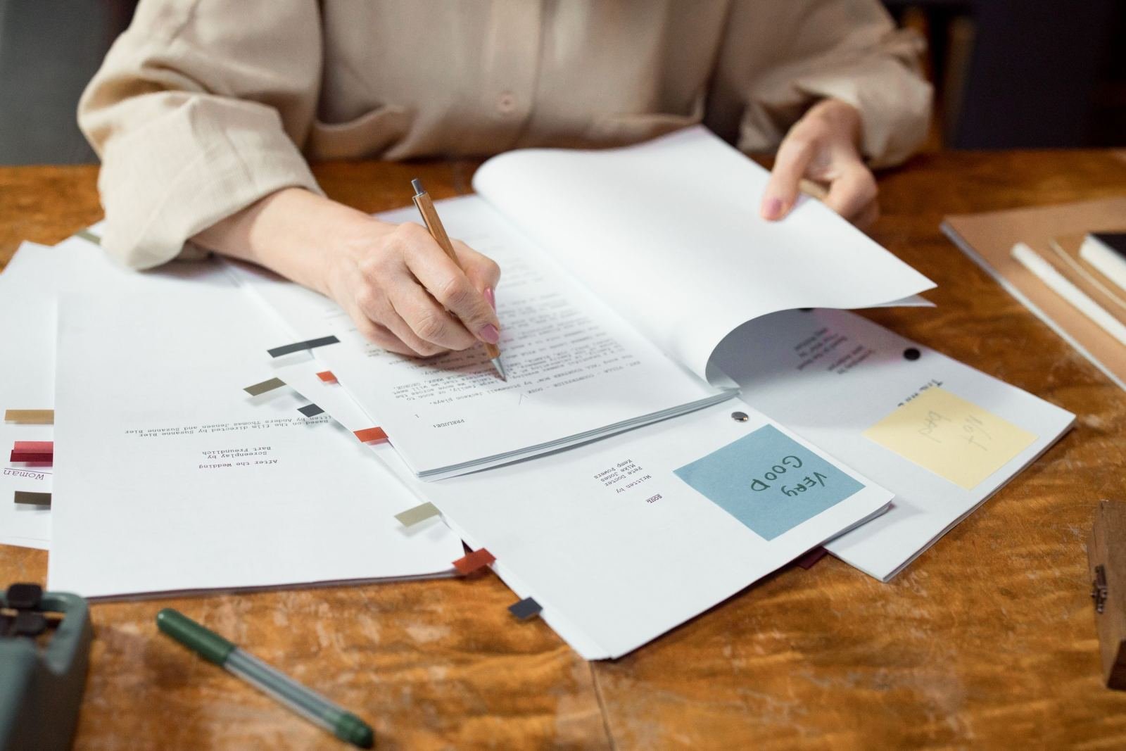 A person reviews notes and a book with colorful sticky tabs on a wooden table