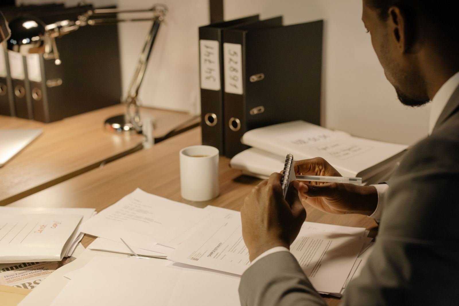 A man in a suit takes notes at a desk with papers and binders
