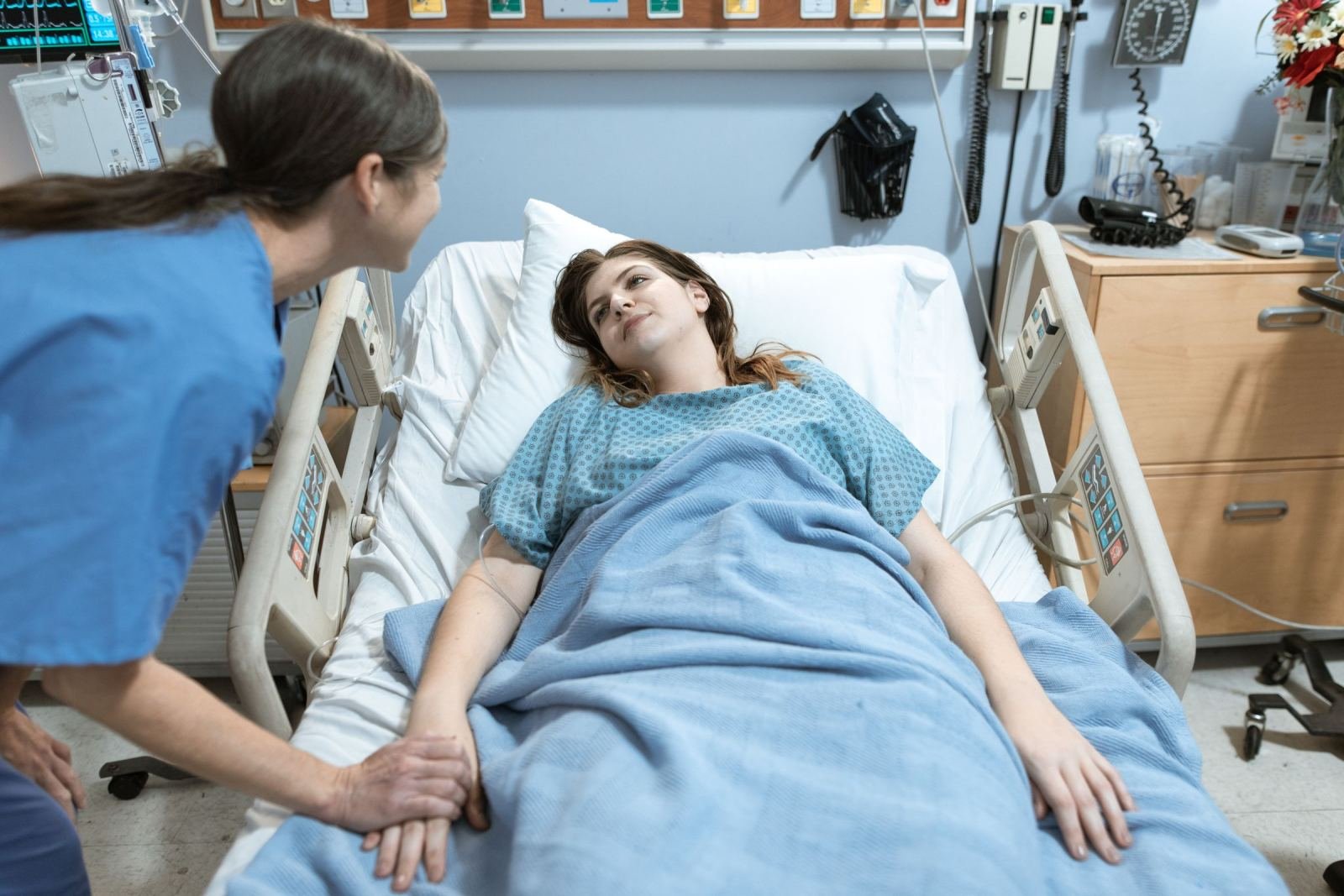 A nurse in blue scrubs holds the hand of a patient lying in a hospital bed