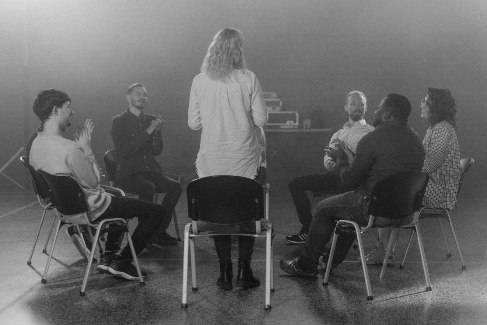 A group of people sitting in a circle of chairs listening to a person standing in the center