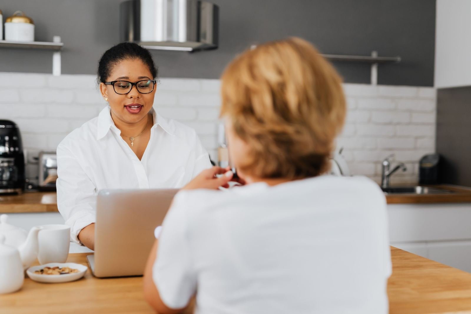 Two women talking at a kitchen counter with a laptop and mugs