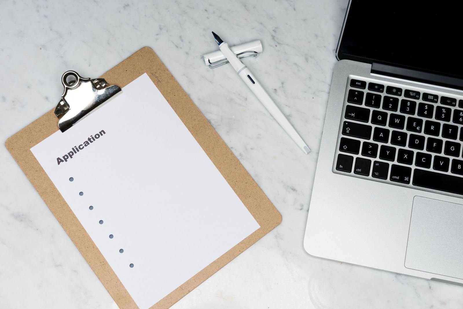 A clipboard with an application form, a pen, and a laptop on a marble surface