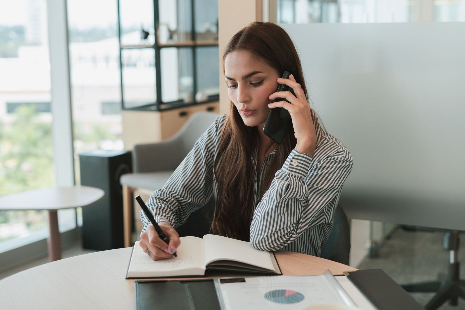 A woman talks on a phone while writing notes in a notebook at a table